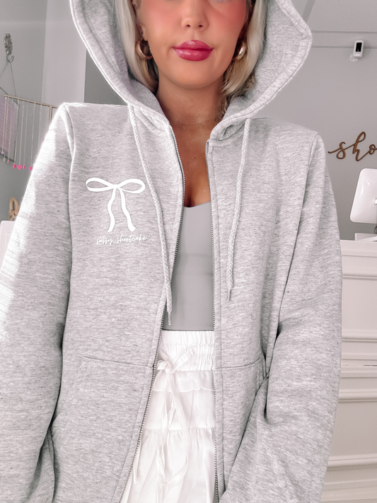 Grey zip-up hoodie featuring a bow and text detail, shown on a model.