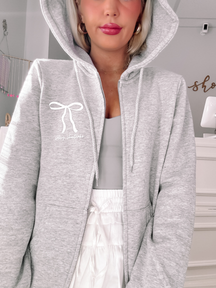 Grey zip-up hoodie featuring a bow and text detail, shown on a model.
