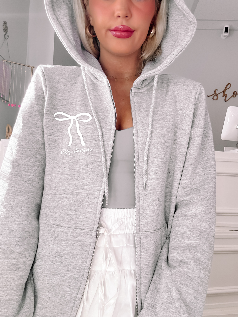 Grey zip-up hoodie featuring a bow and text detail, shown on a model.
