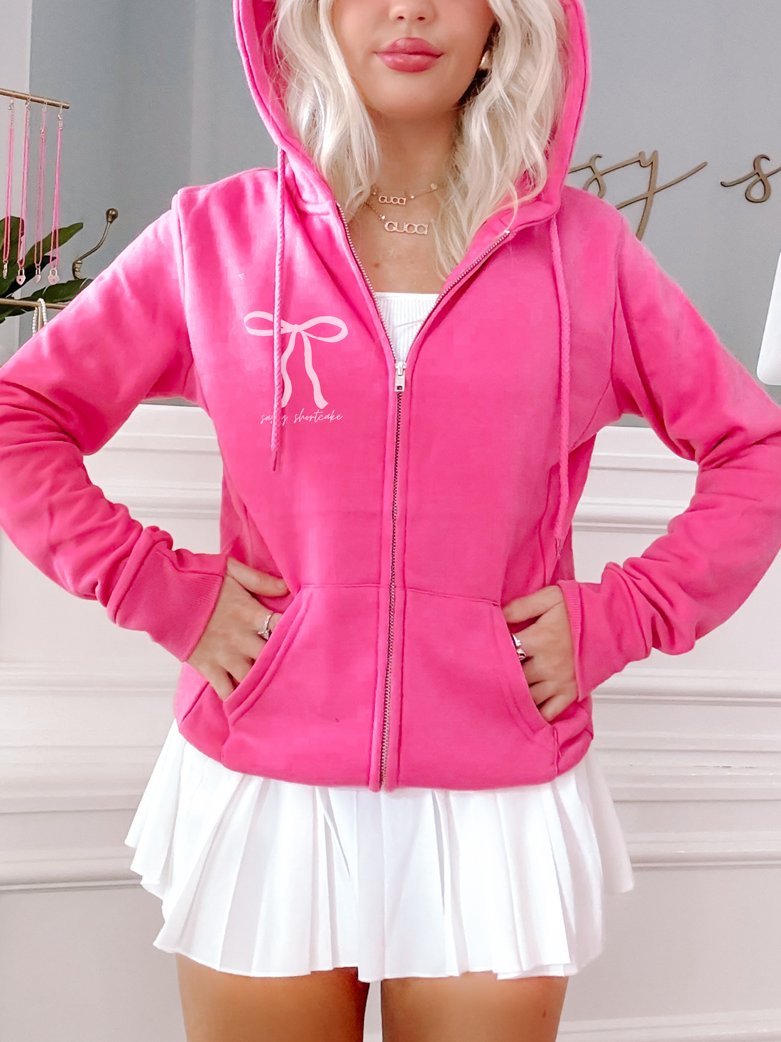 Hot pink zip-up hoodie featuring a bow graphic, modeled with a white pleated skirt.
