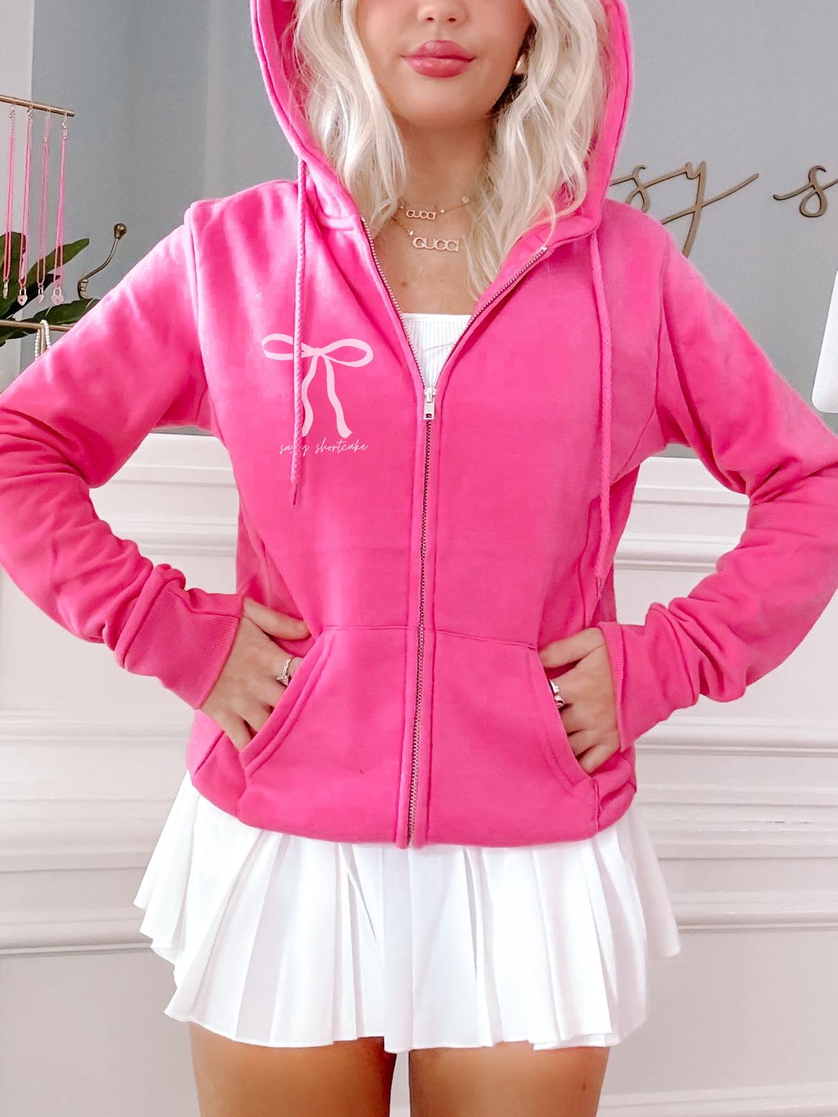 Hot pink zip-up hoodie featuring a bow design, modeled with a white pleated skirt.
