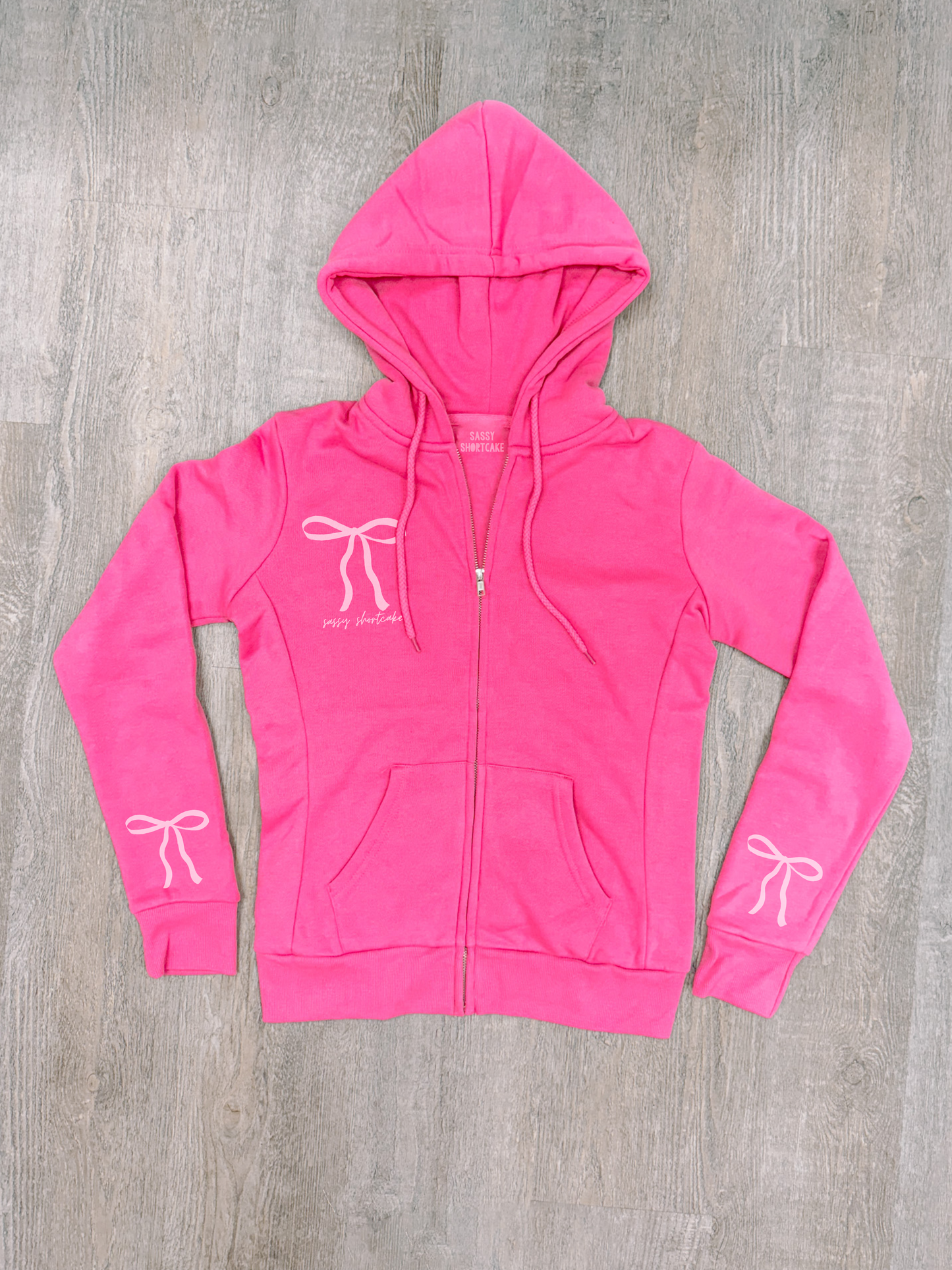 Hot pink zip-up hoodie with bow details, shown on wood background.
