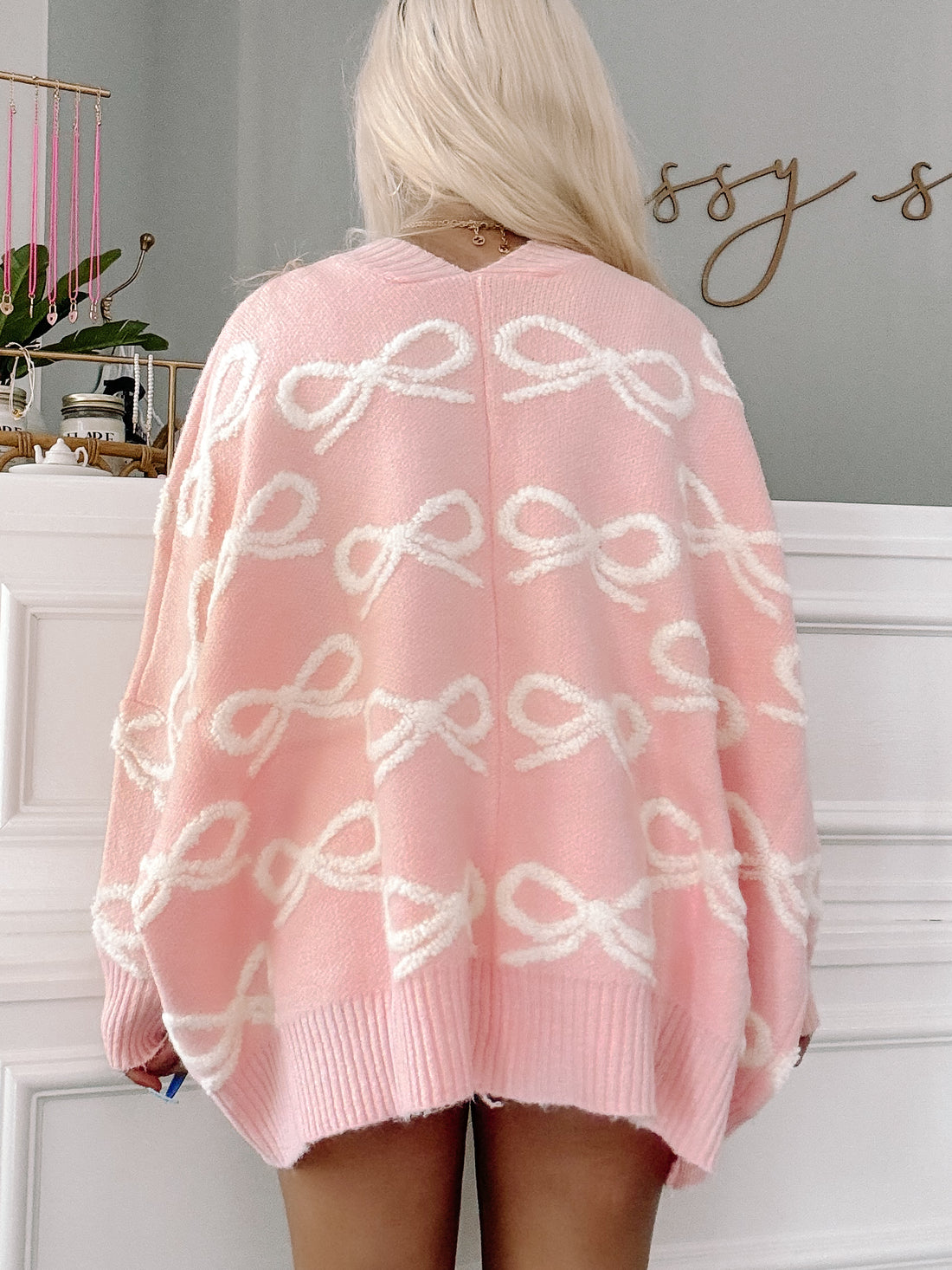 Pink knit cardigan featuring a white bow pattern, shown from the back against a white wall | Sassy Shortcake
