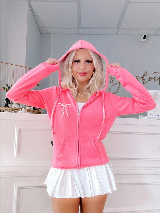 Neon pink zip-up hoodie with bow detail, modeled against a white wall.
