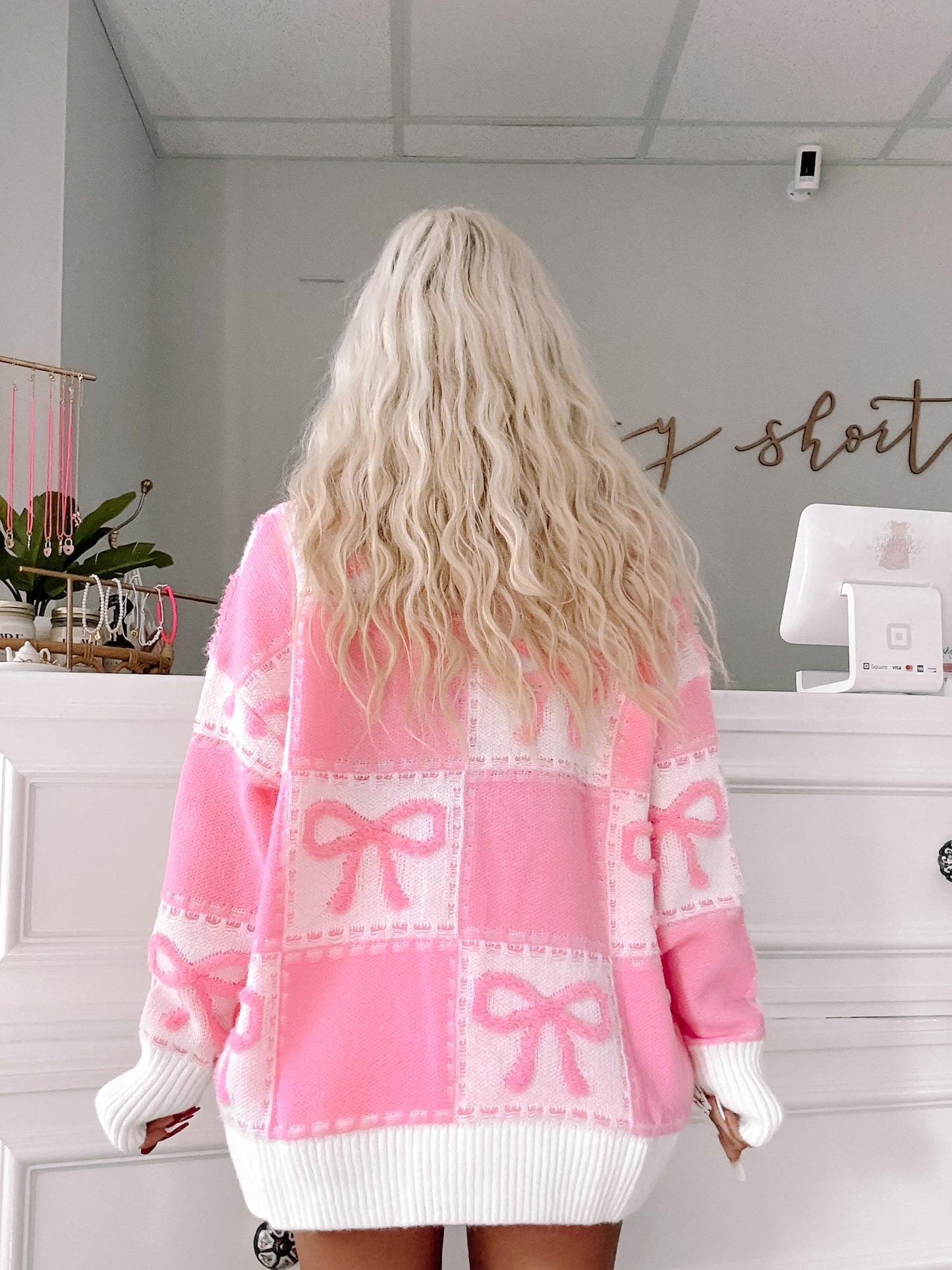 Pink knit sweater featuring a playful bow pattern, shown from the back in a boutique setting.
