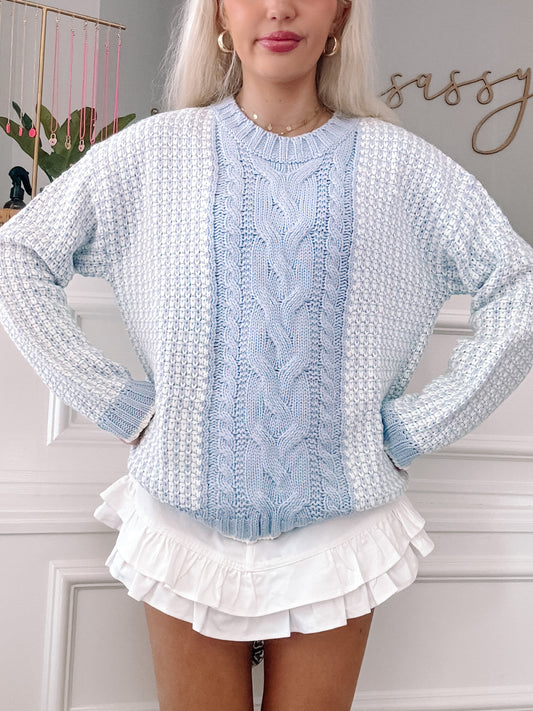 Light blue cable-knit sweater with textured detailing, styled with a white ruffled skirt.