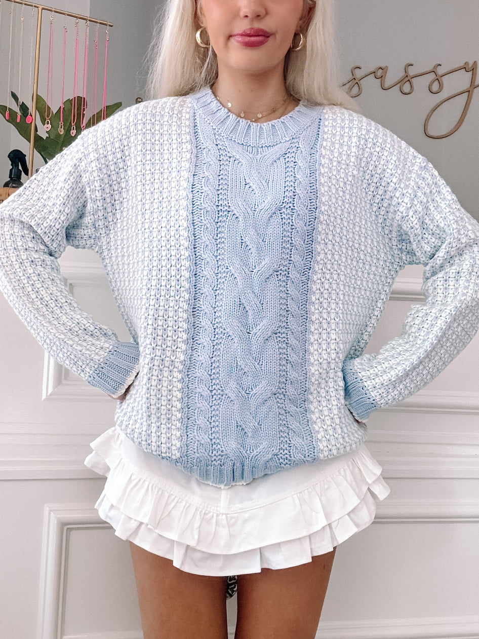 Light blue cable-knit sweater with textured detailing, styled with a white ruffled skirt.
