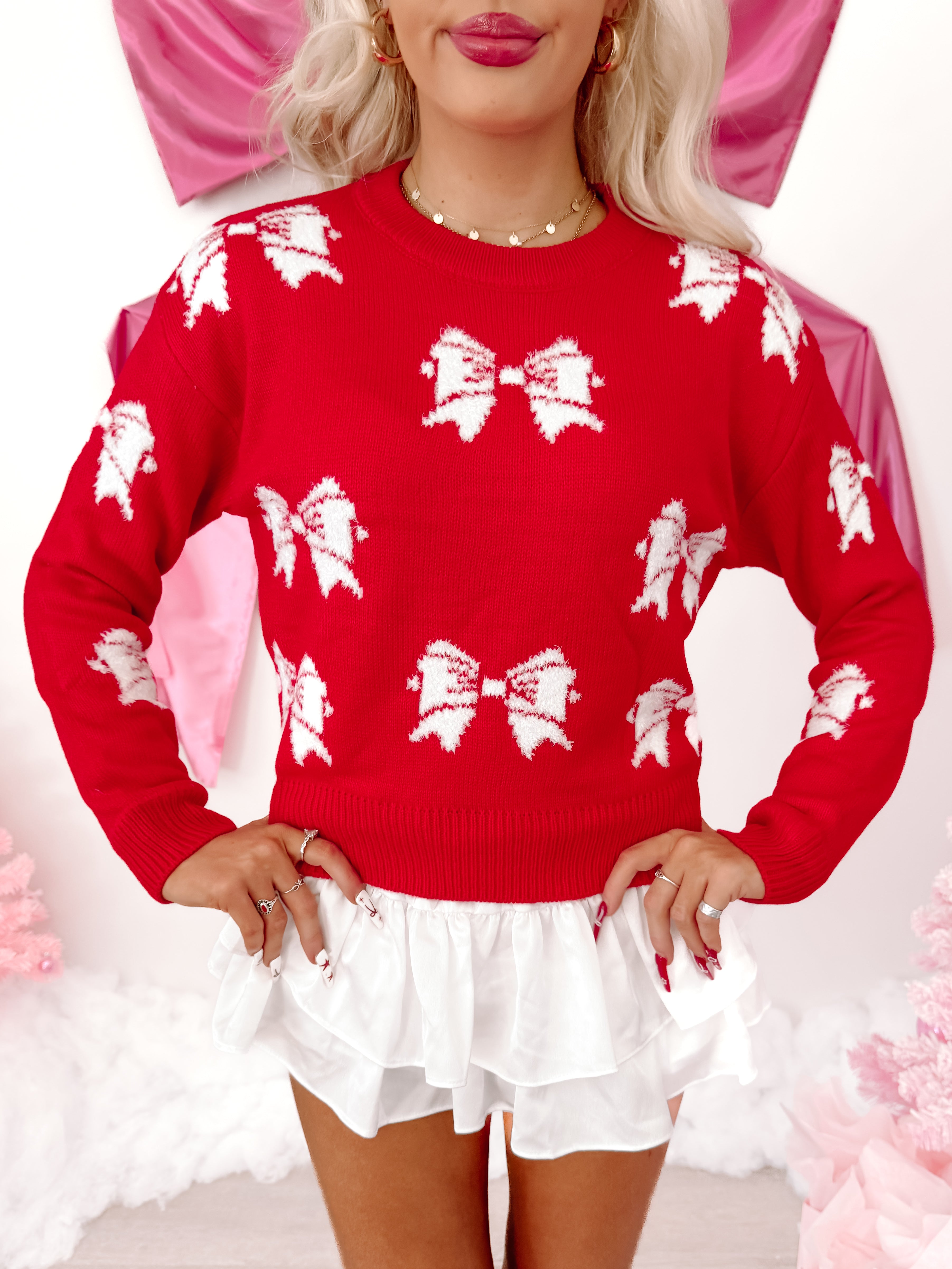 Red knit sweater with white bow pattern, modeled against a pink backdrop.
