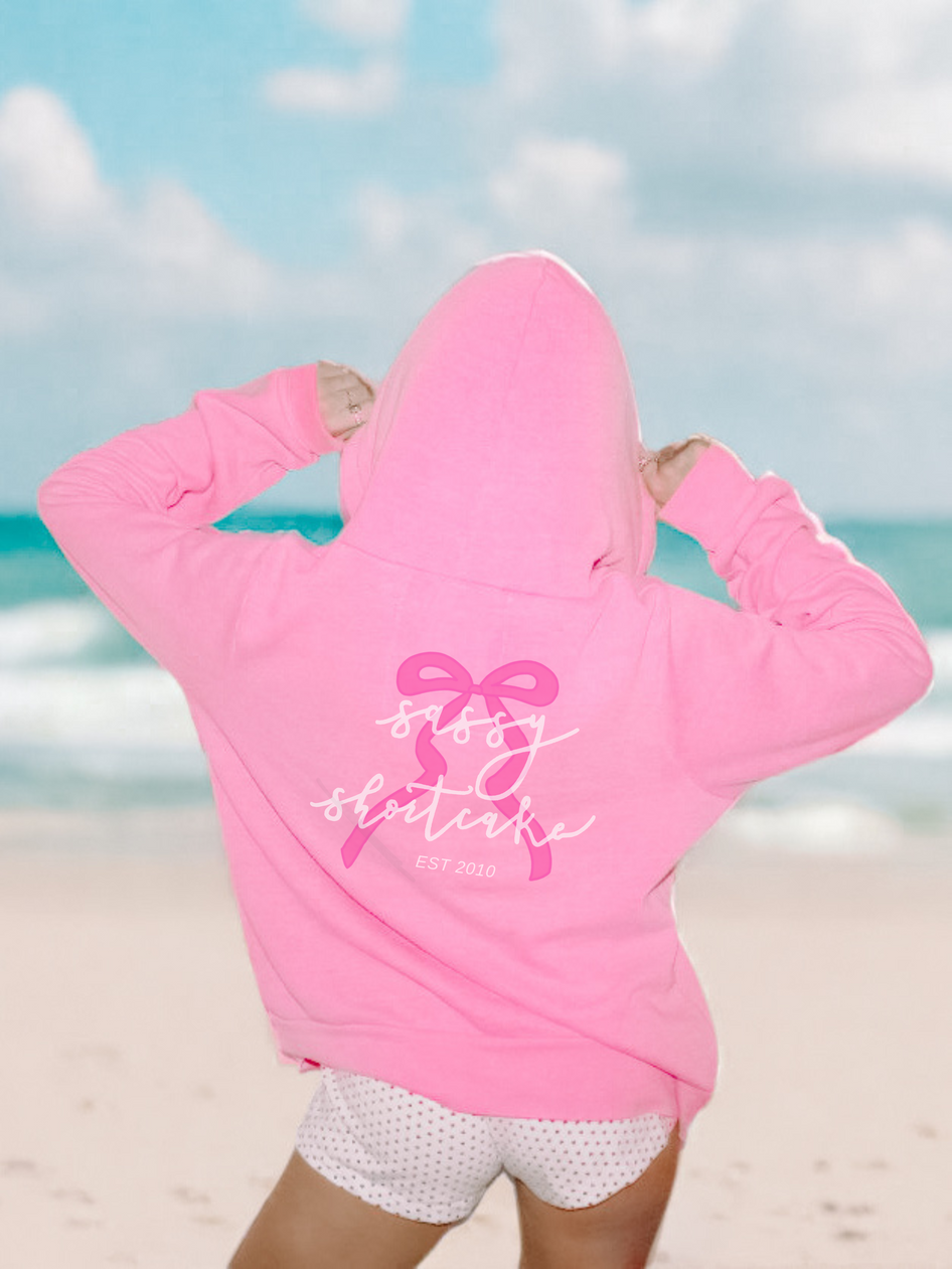 Pink hoodie on beach background, featuring a pink bow and script logo.
