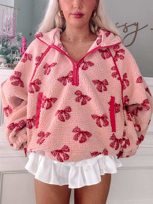 Pink sherpa pullover with red bow print, half-zipped, modeled over a white ruffle skirt.