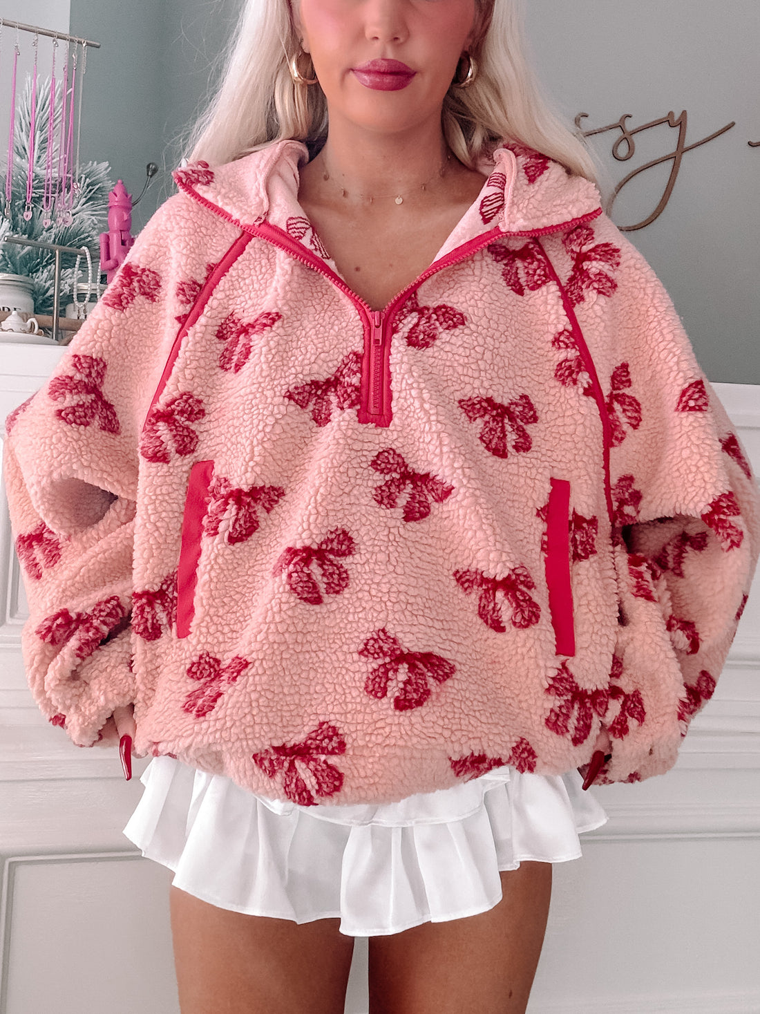 Pink sherpa pullover with red bow print, half-zipped, modeled over a white ruffle skirt.
