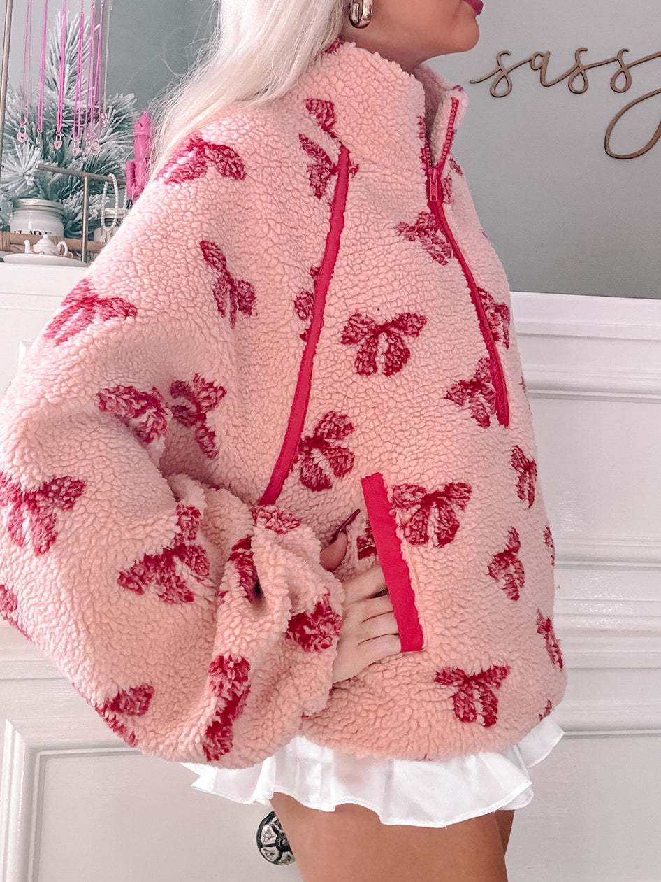 Pink sherpa pullover with red bow pattern, half-zip closure.
