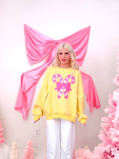 Model wearing a bright yellow crewneck with a festive, sparkly sequin bow and candy cane graphic on the front from Sassy Shortcake