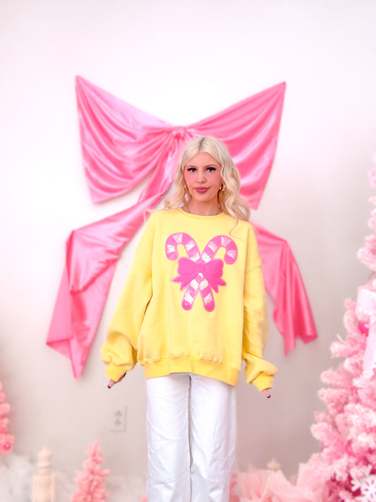 Model wearing a bright yellow crewneck with a festive, sparkly sequin bow and candy cane graphic on the front from Sassy Shortcake