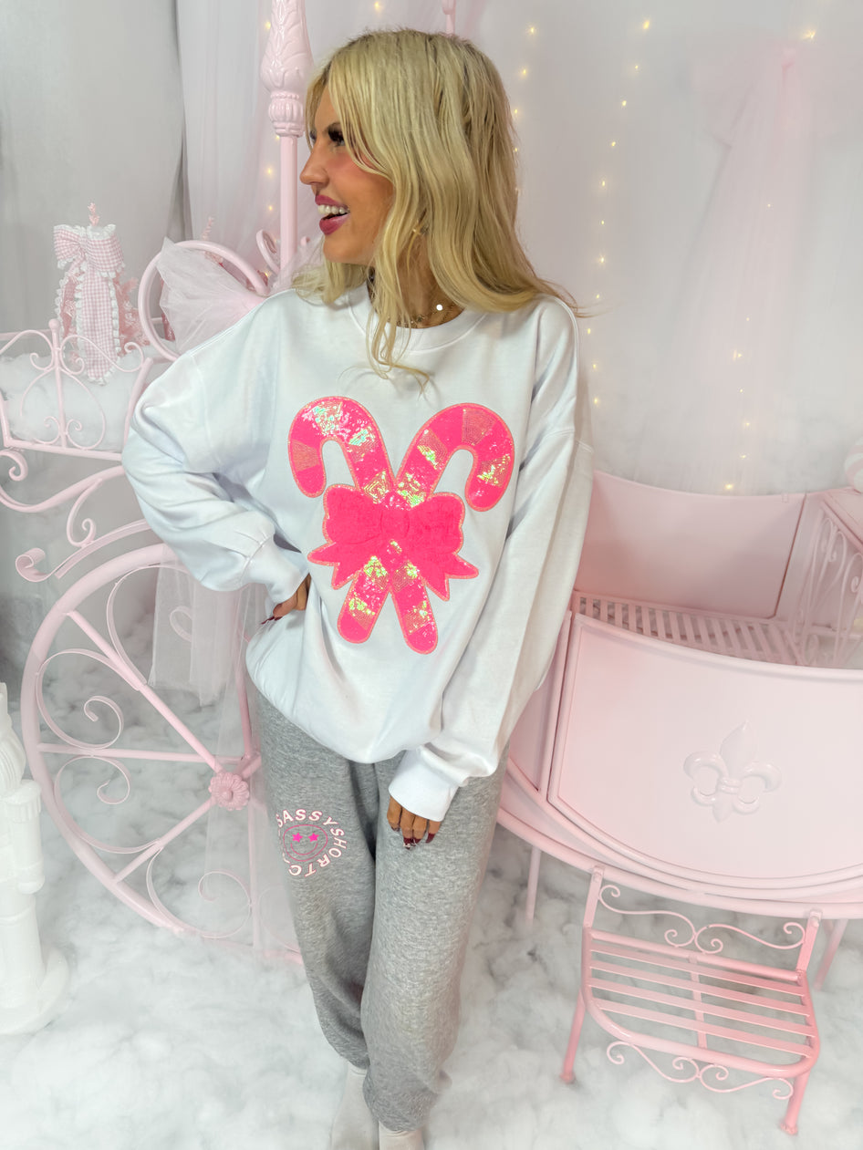 White crewneck sweatshirt featuring pink sequined candy canes, modeled in a pastel pink room.
