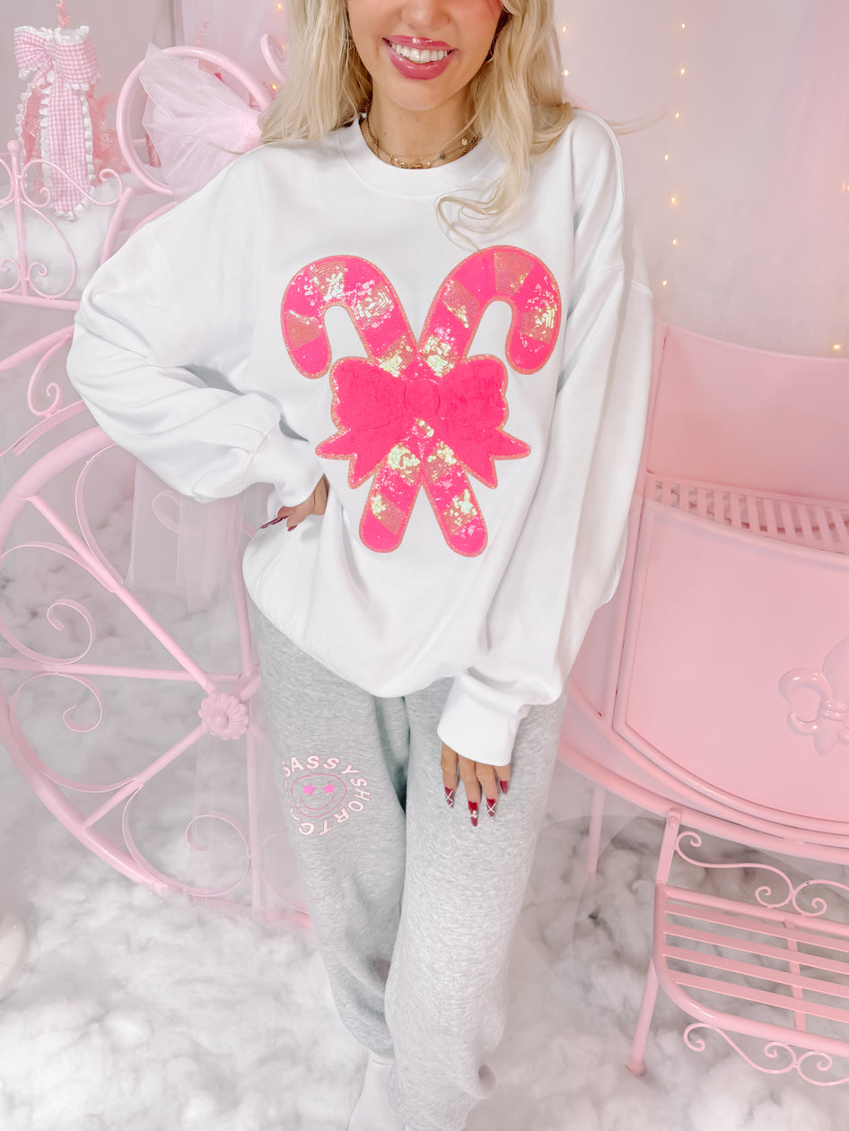 White crewneck sweatshirt featuring pink sequined candy canes and bow; festive apparel.

