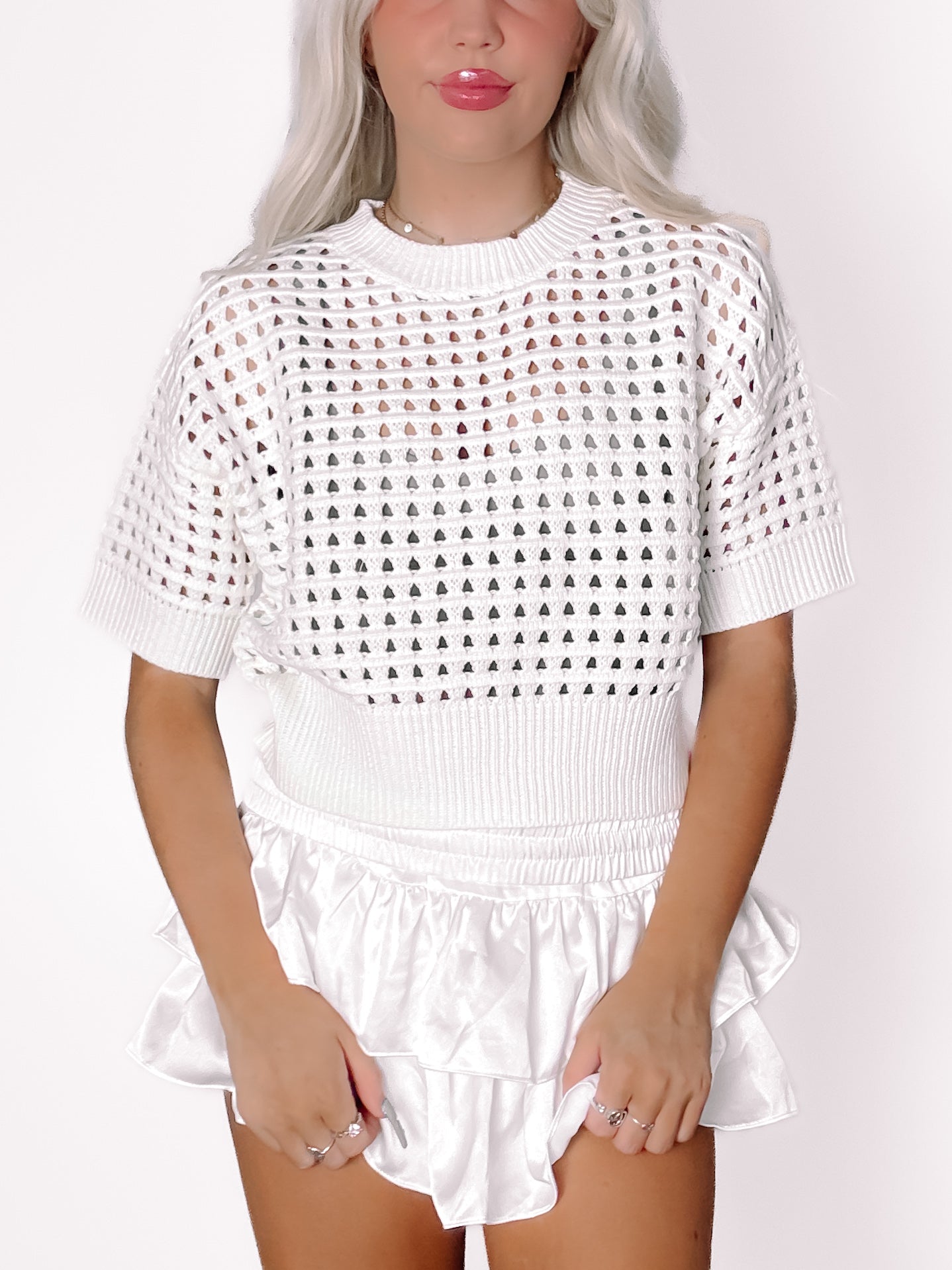 White crochet-knit top with short sleeves and eyelet detailing, styled with a white satin skirt.
