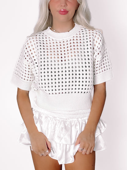 White crochet-knit top with short sleeves and eyelet detailing, styled with a white satin skirt.
