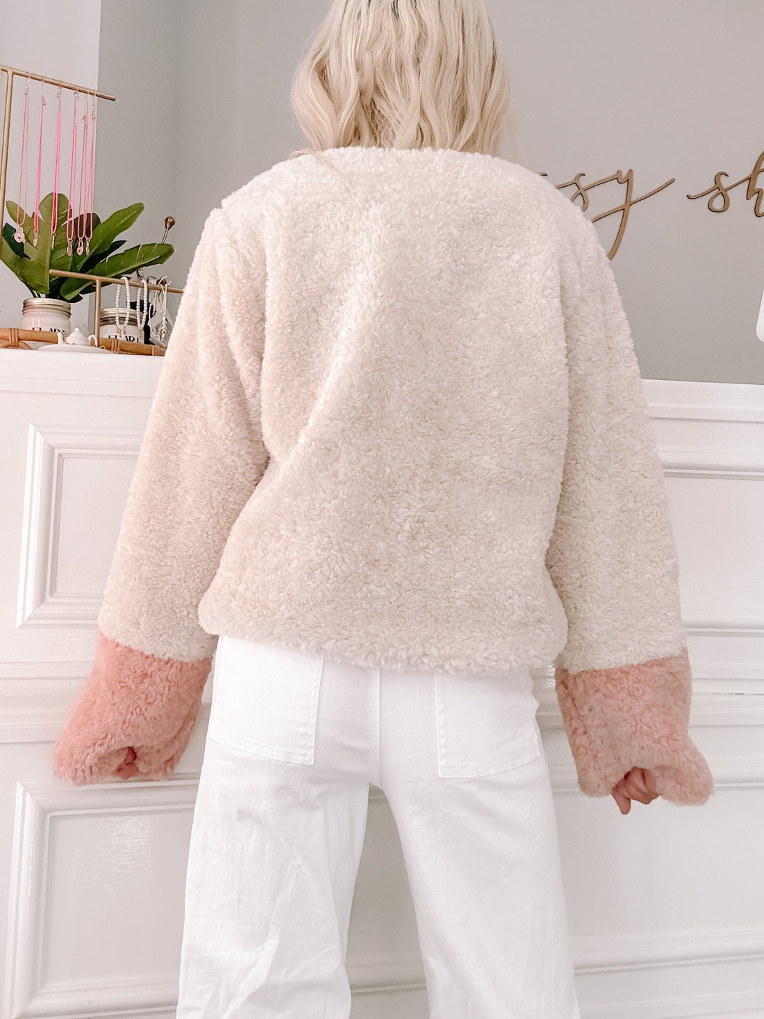 Cream sherpa jacket with blush pink cuffs, shown from the back against a white wall.
