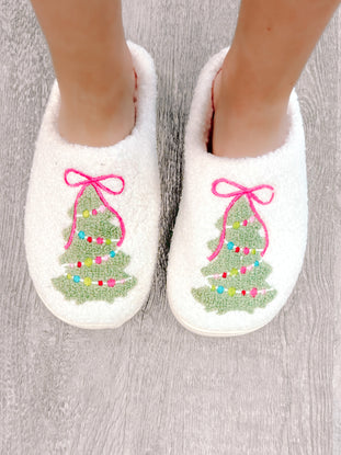 Cozy white slippers adorned with embroidered Christmas trees and pink bows, shown on a wood floor.
