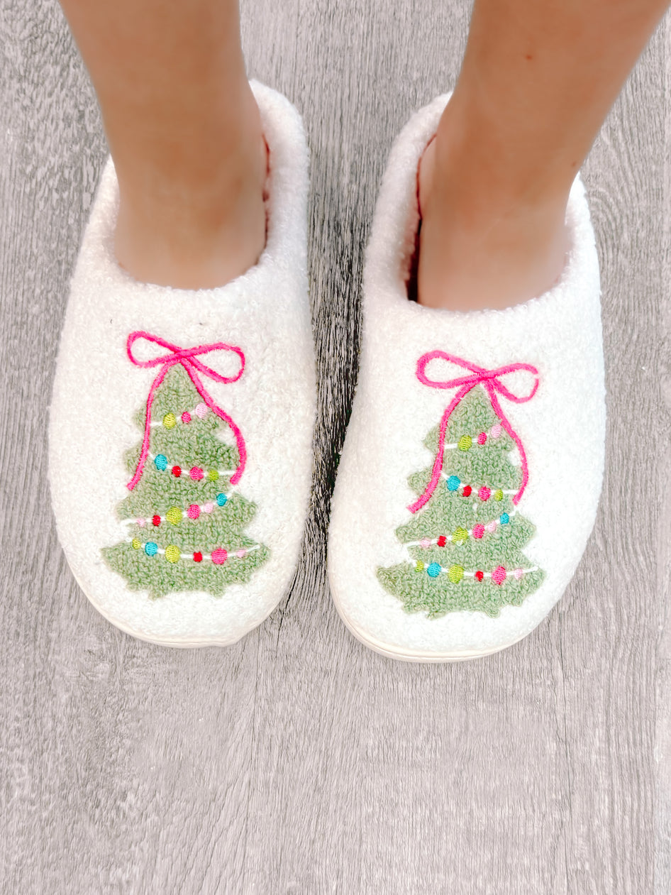 Cozy white slippers adorned with embroidered Christmas trees and pink bows, shown on a wood floor.
