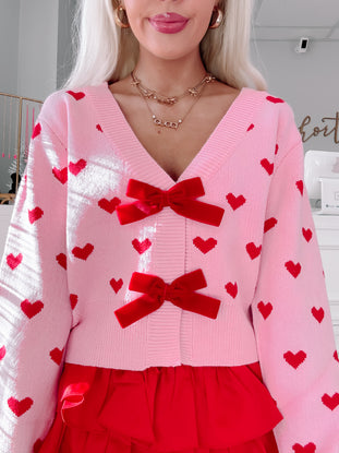Pink knit cardigan adorned with red hearts and velvet bows, styled with a red ruffled skirt.
