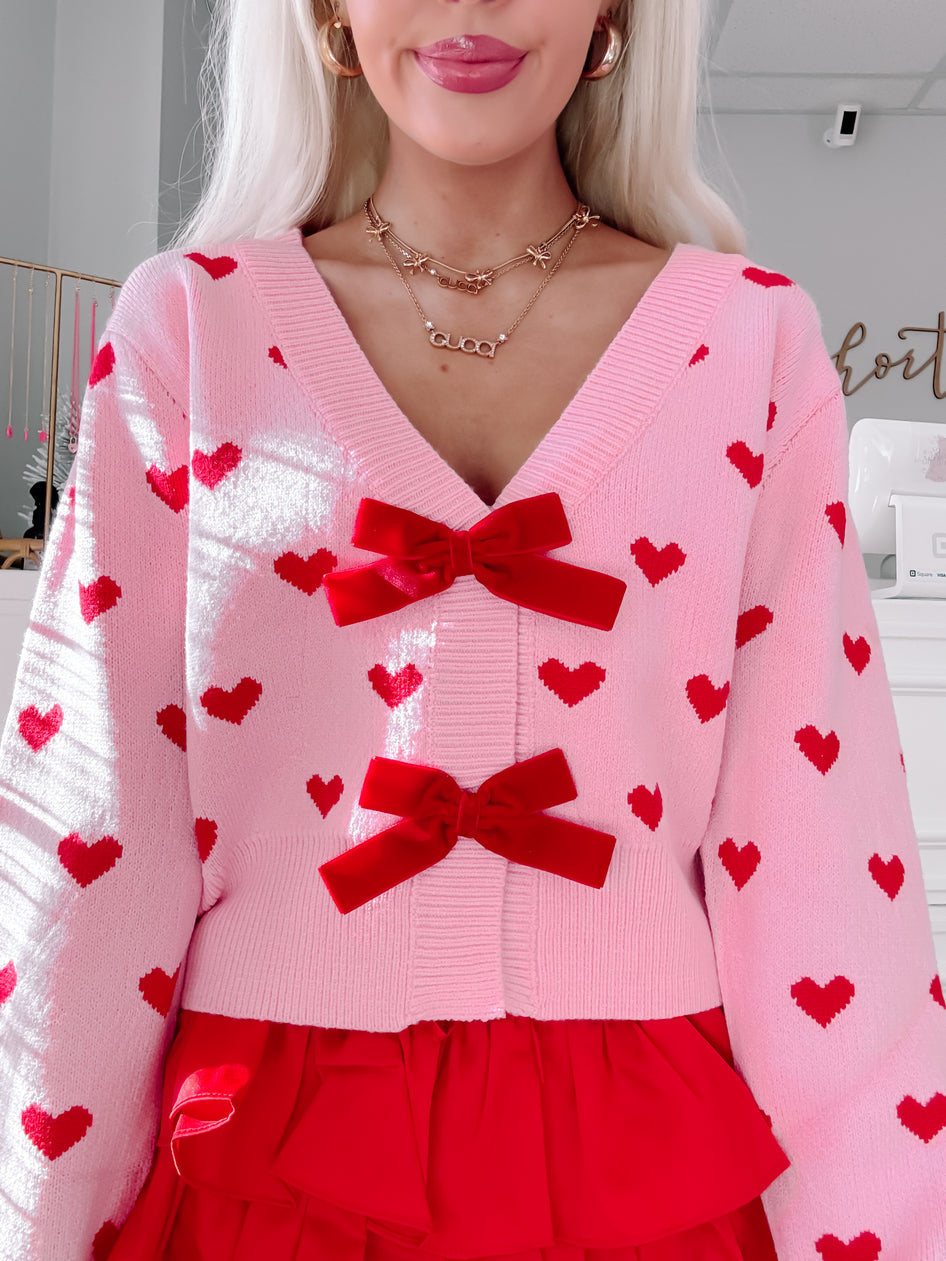 Pink knit cardigan adorned with red hearts and velvet bows, styled with a red ruffled skirt.
