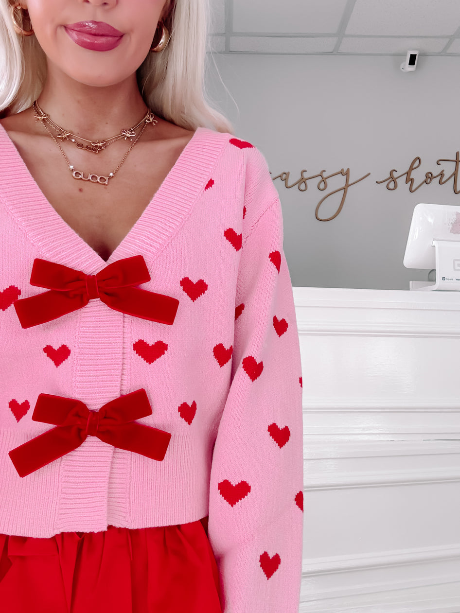 Pink knit cardigan adorned with red hearts and velvet bows, shown on a model.
