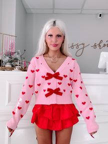 Pink knit cardigan with red heart pattern and velvet bows, modeled indoors.
