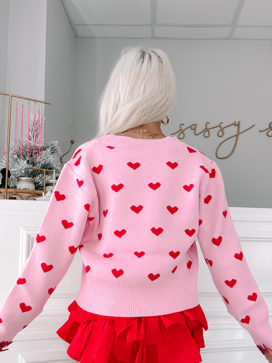 Pink knit cardigan adorned with small red hearts, shown from the back against a light-grey wall.
