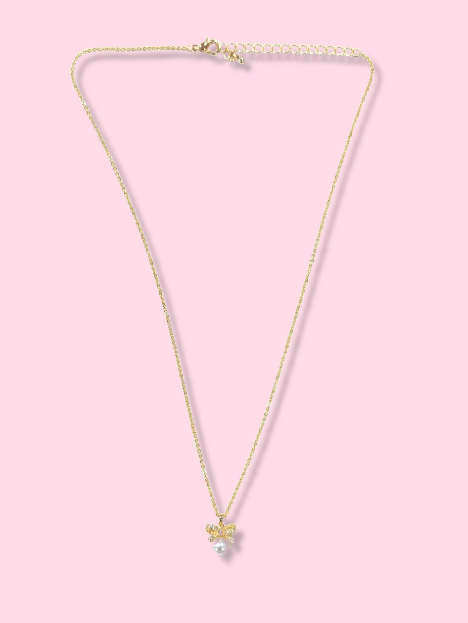 Gold chain necklace with a dainty bow and pearl pendant, displayed against a pink background.
