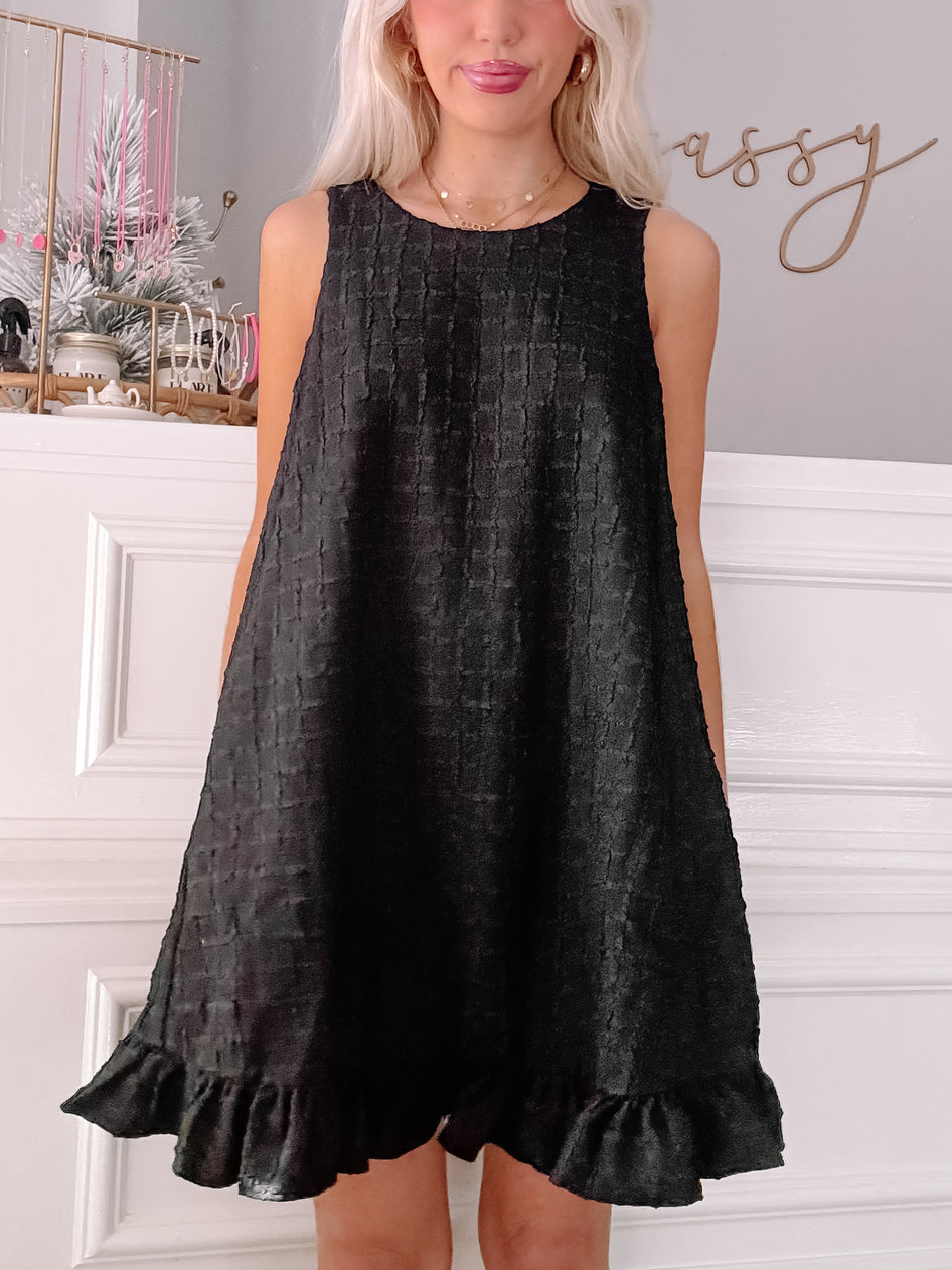 Black sleeveless mini dress with textured fabric and ruffled hem, modeled against a white wall.
