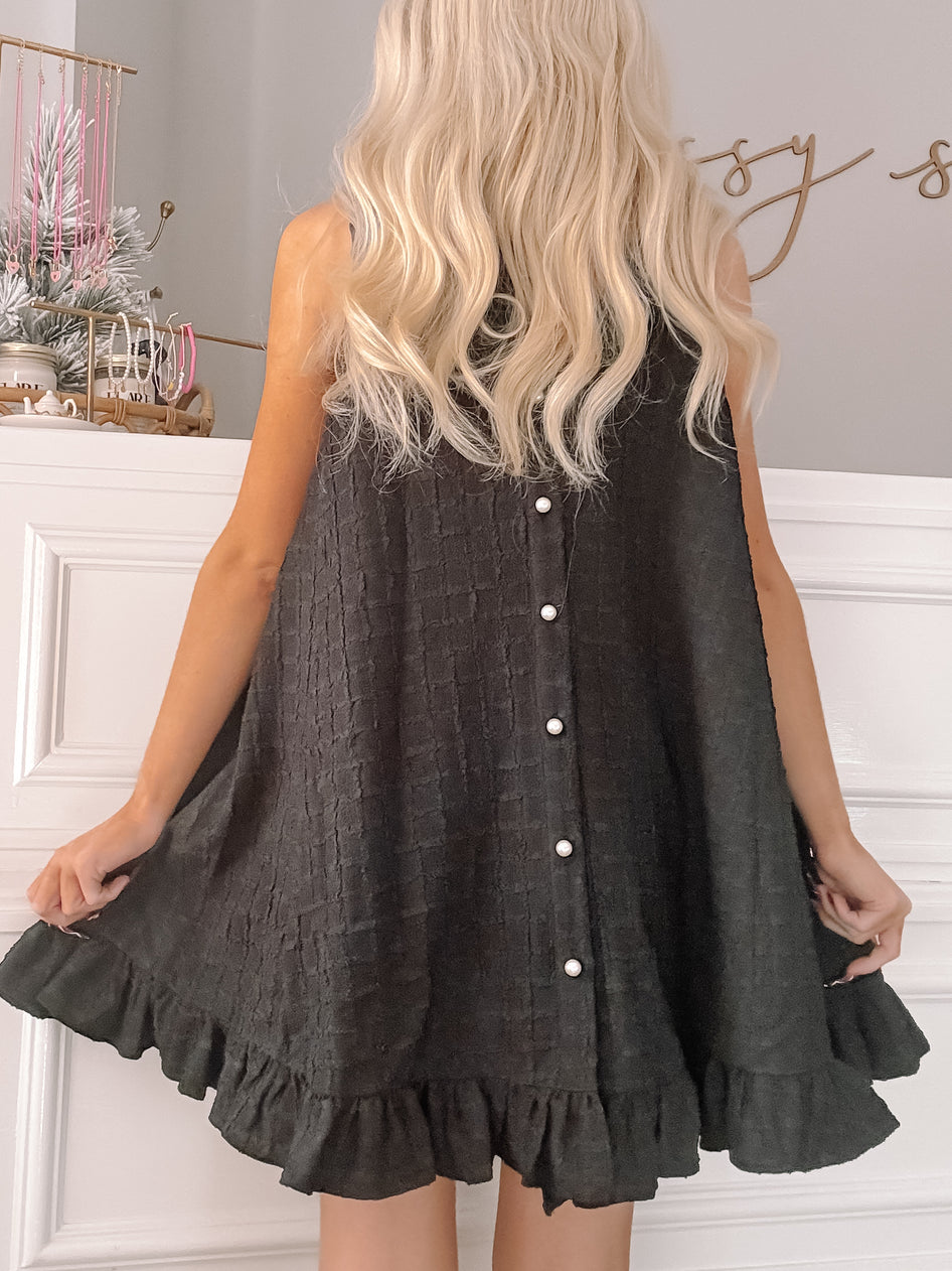 Black sleeveless mini dress with a textured bodice, pearl buttons, and ruffled hem, shown from the back.
