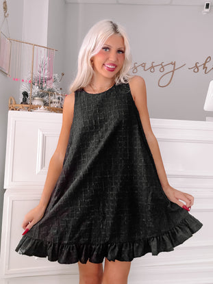 Black sleeveless mini dress with textured fabric and ruffled hem, modeled in a boutique.
