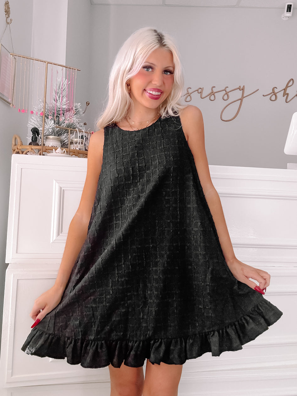 Black sleeveless mini dress with textured fabric and ruffled hem, modeled in a boutique.
