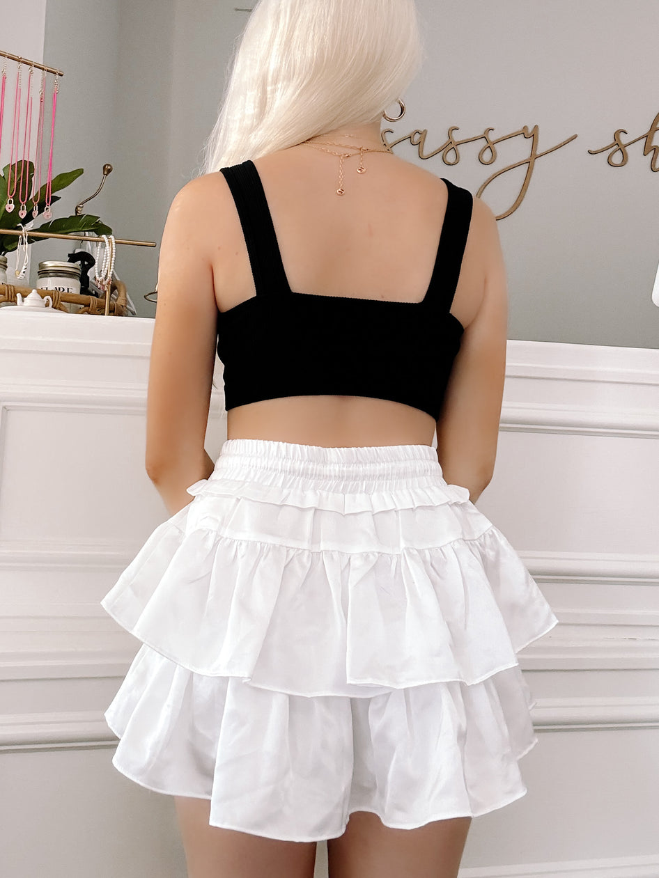 Rear view of a woman wearing a black cropped tank top and a white tiered ruffle mini skirt against a neutral backdrop.
