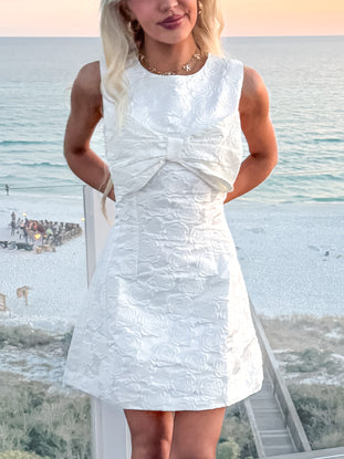 White rose-embossed dress with a bow detail, shown outdoors near the ocean.
