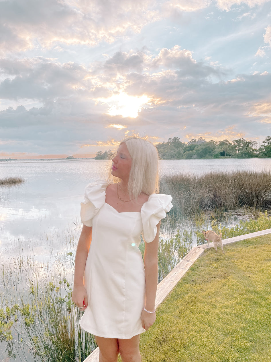 White puff-sleeve mini dress by the water at sunset.
