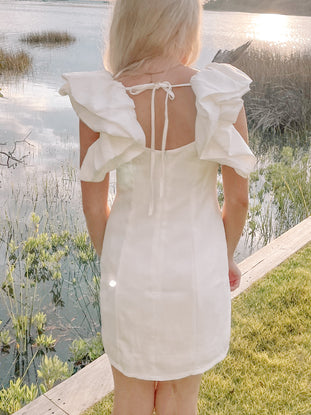 White mini dress with ruffled sleeves, viewed from behind by a body of water.
