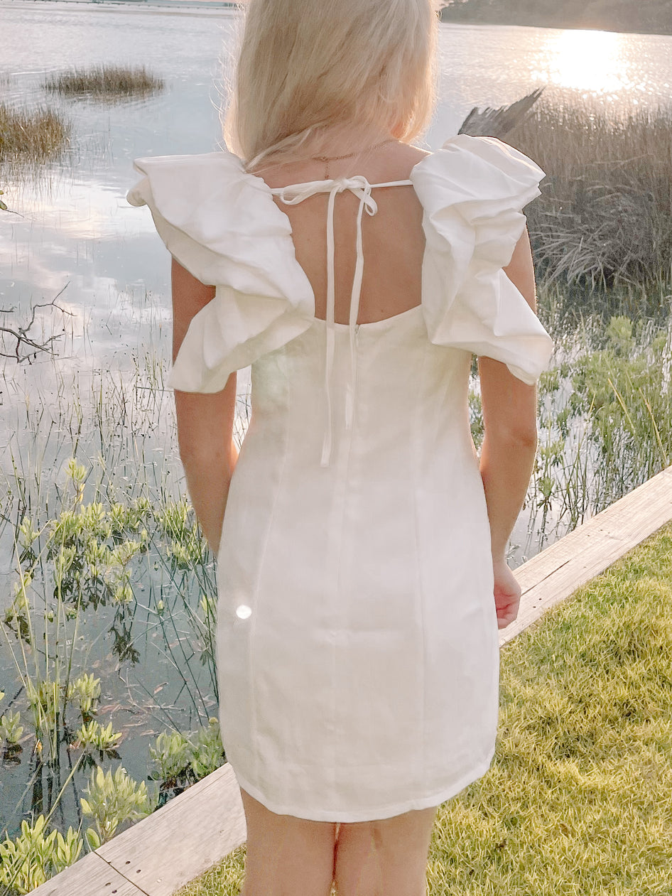 White mini dress with ruffled sleeves, viewed from behind by a body of water.
