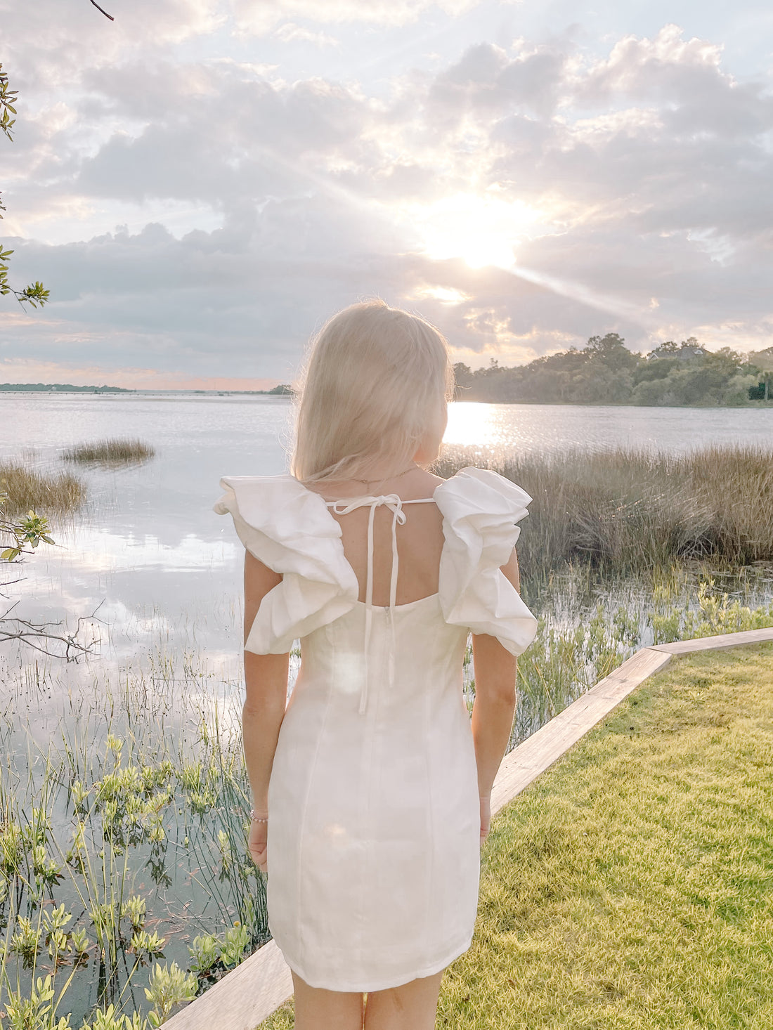White mini dress with ruffled sleeves, viewed from behind against a tranquil sunset lakeside scene.
