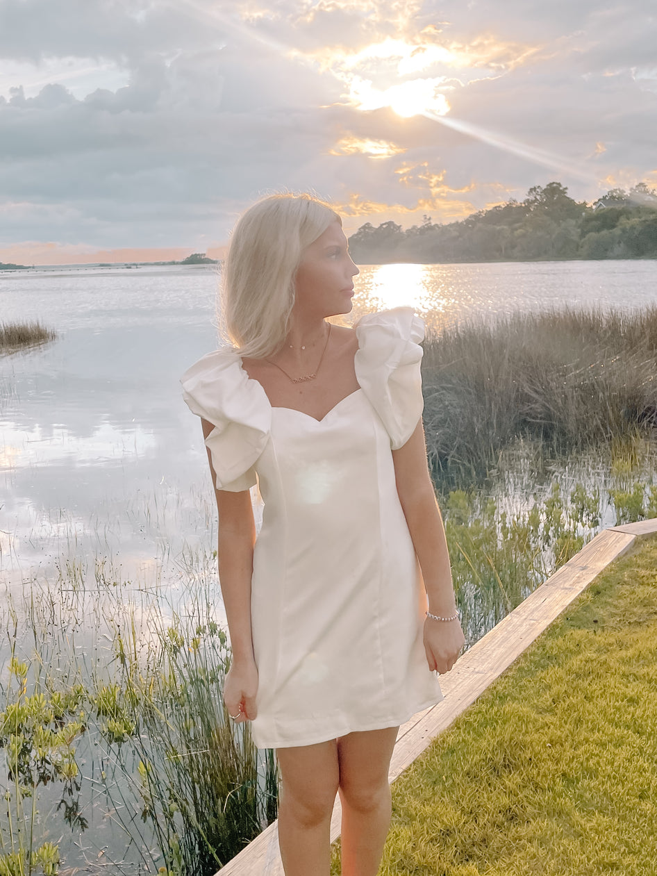 White puff-sleeve mini dress by the water at sunset.
