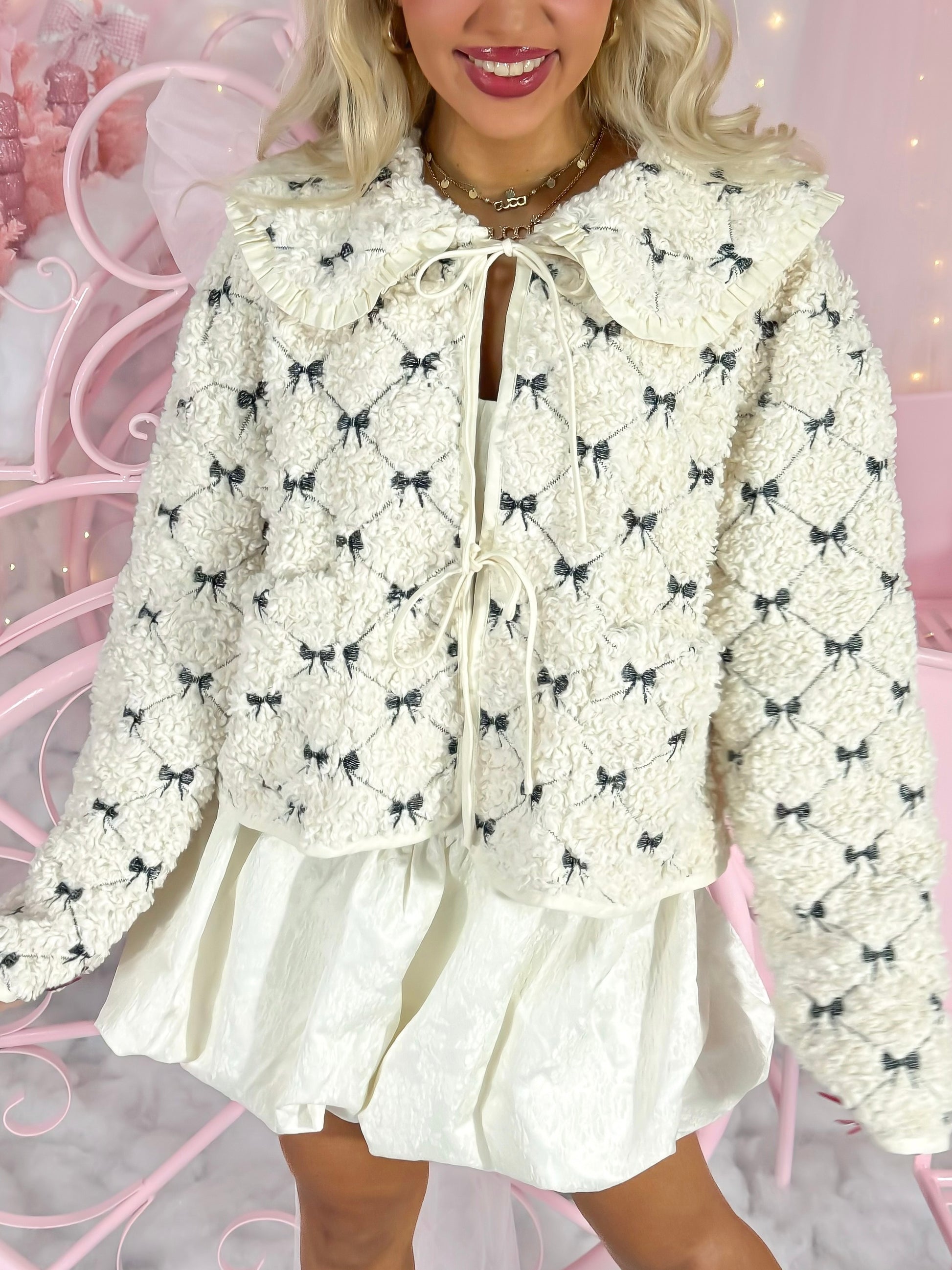 Cream-colored fluffy jacket with dark bows, featuring a ruffled collar and tie closure.
