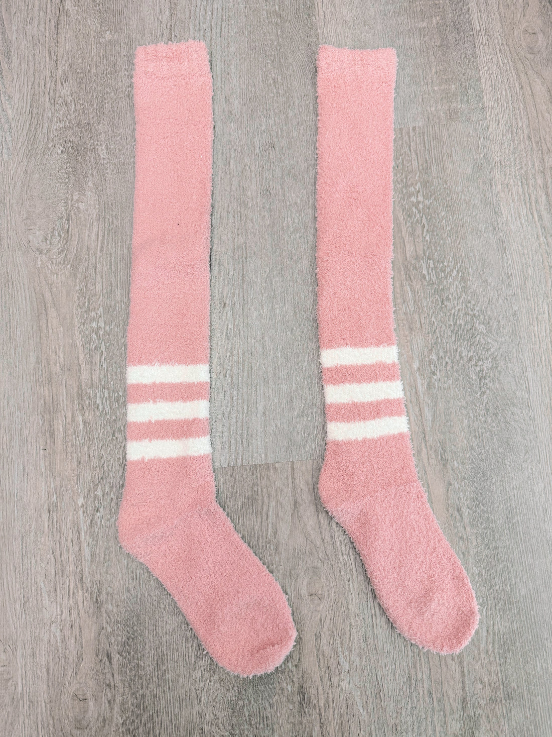 Pair of fluffy pink knee-high socks with white stripes, displayed on wood.
