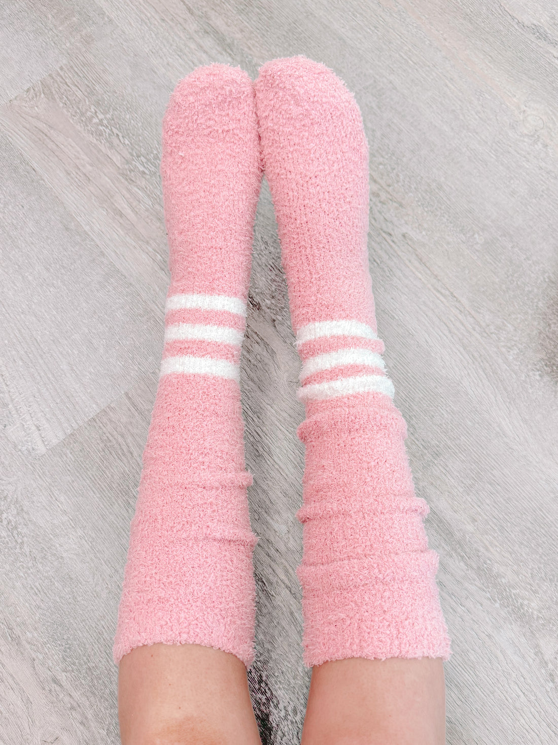 Pink fuzzy socks with white stripes, cozy and comfy, shown on legs against a wood floor.
