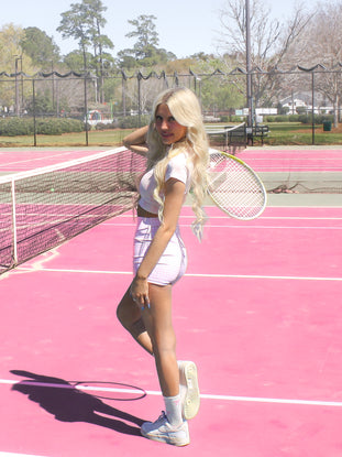 Woman in gingham shorts and tennis shoes on a pink court with a tennis racket.
