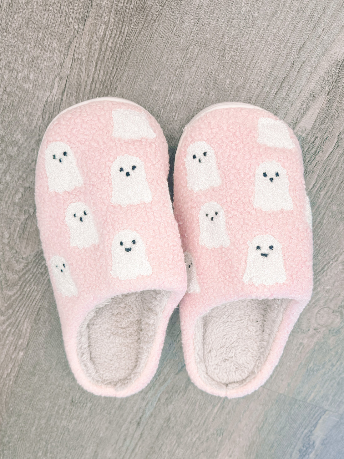 Pink fuzzy slippers with a whimsical ghost pattern, shown on a wood floor.
