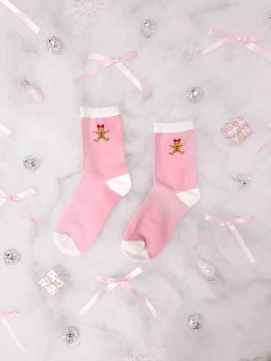 Pink crew socks with gingerbread man design, nestled amongst festive decorations on marble.