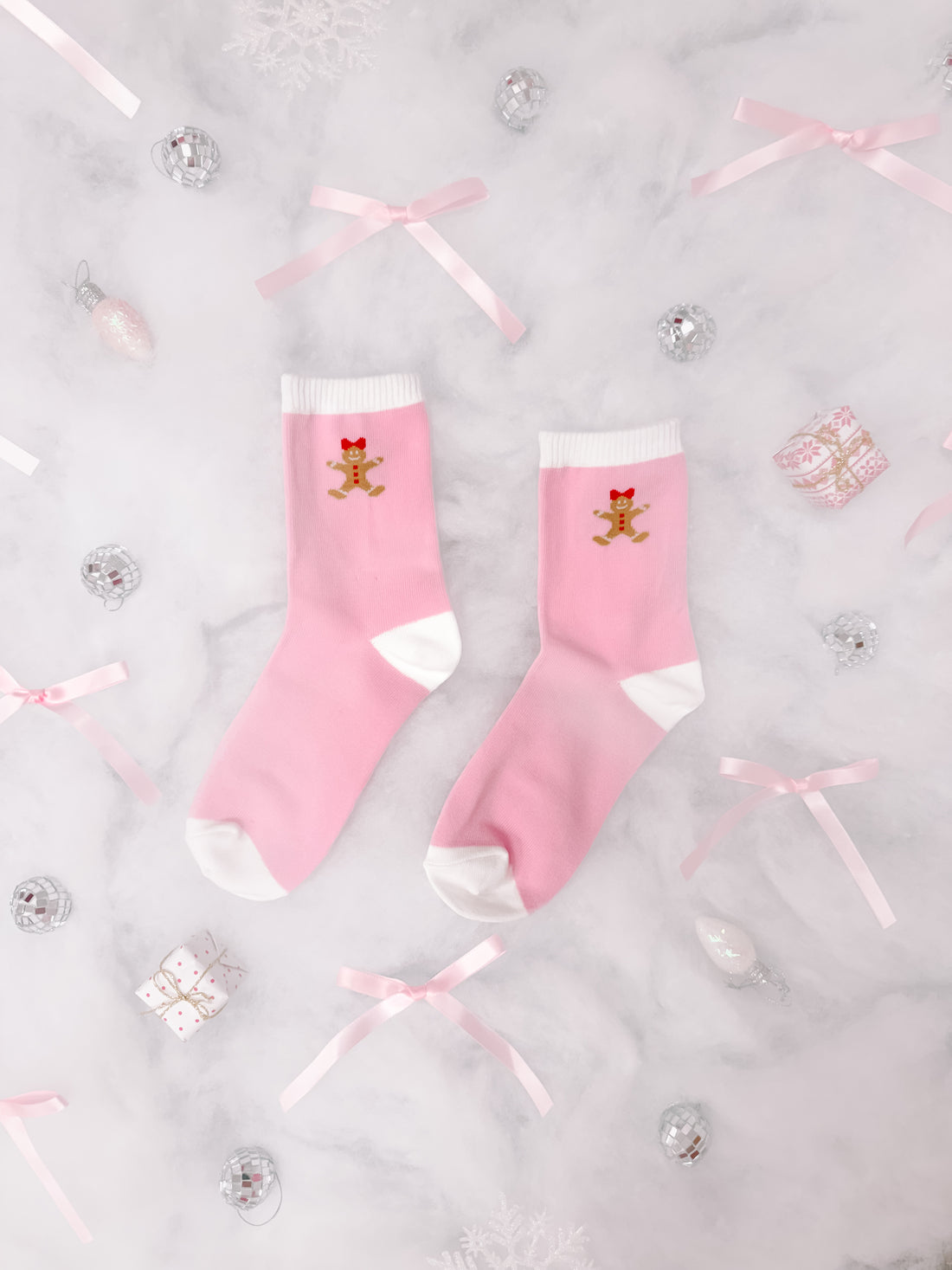Pink crew socks with gingerbread man design, nestled amongst festive decorations on marble.

