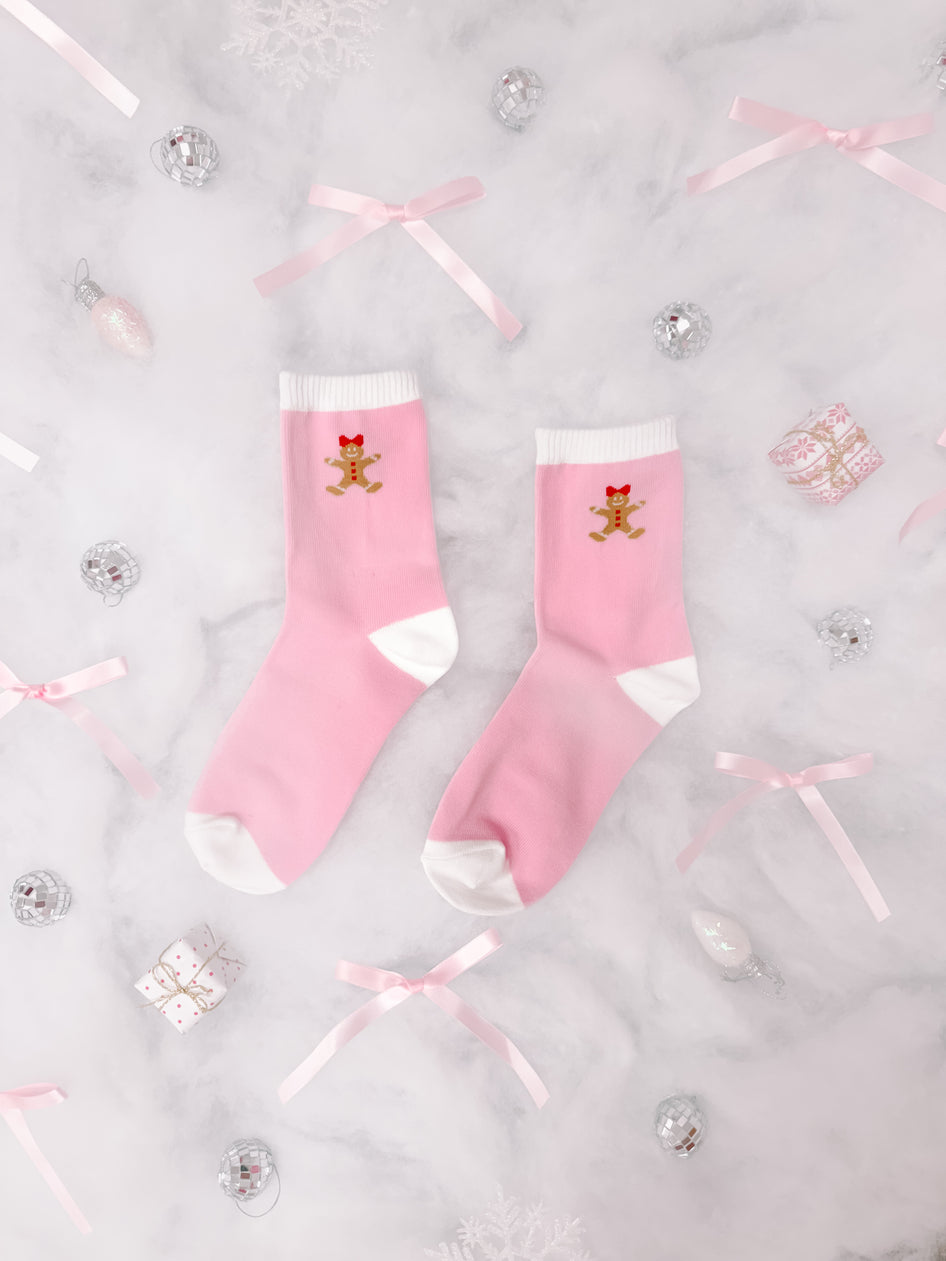 Pink crew socks with gingerbread man design, nestled amongst festive decorations on marble.
